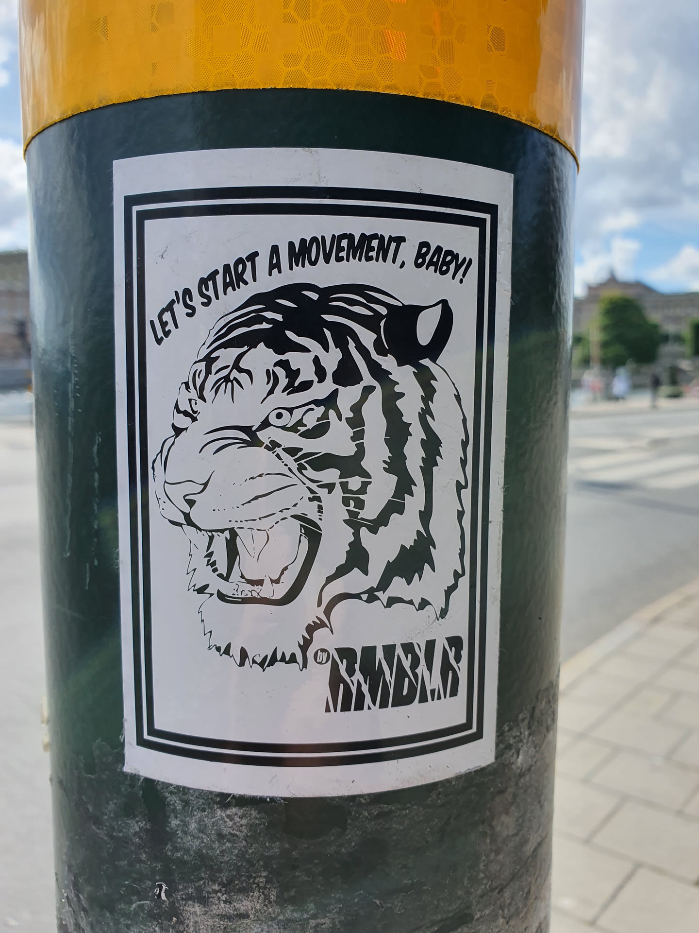 A black and white illustration of a tiger's head with the text "Let's start a movement, baby!" and a logo that says "RMBLR" at the bottom. The sticker is on a dark green pole.