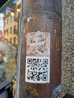 A sticker featuring a QR code and several small polaroid-style photos is affixed to a weathered, rusty metal pole. The photos appear to be of various scenes, though details are indiscernible. The QR code is large and easily scannable. The pole shows signs of age and exposure to the elements.