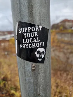 A black and white sticker with a slightly menacing face and the text: "SUPPORT YOUR LOCAL PSYCHO!!!"
