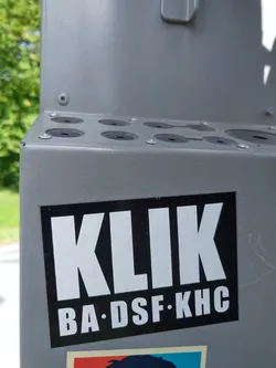 A black and white sticker with the word "KLIK" in large white letters, and underneath in smaller letters "BA·DSF·KHC". It's affixed to a gray metal surface.
