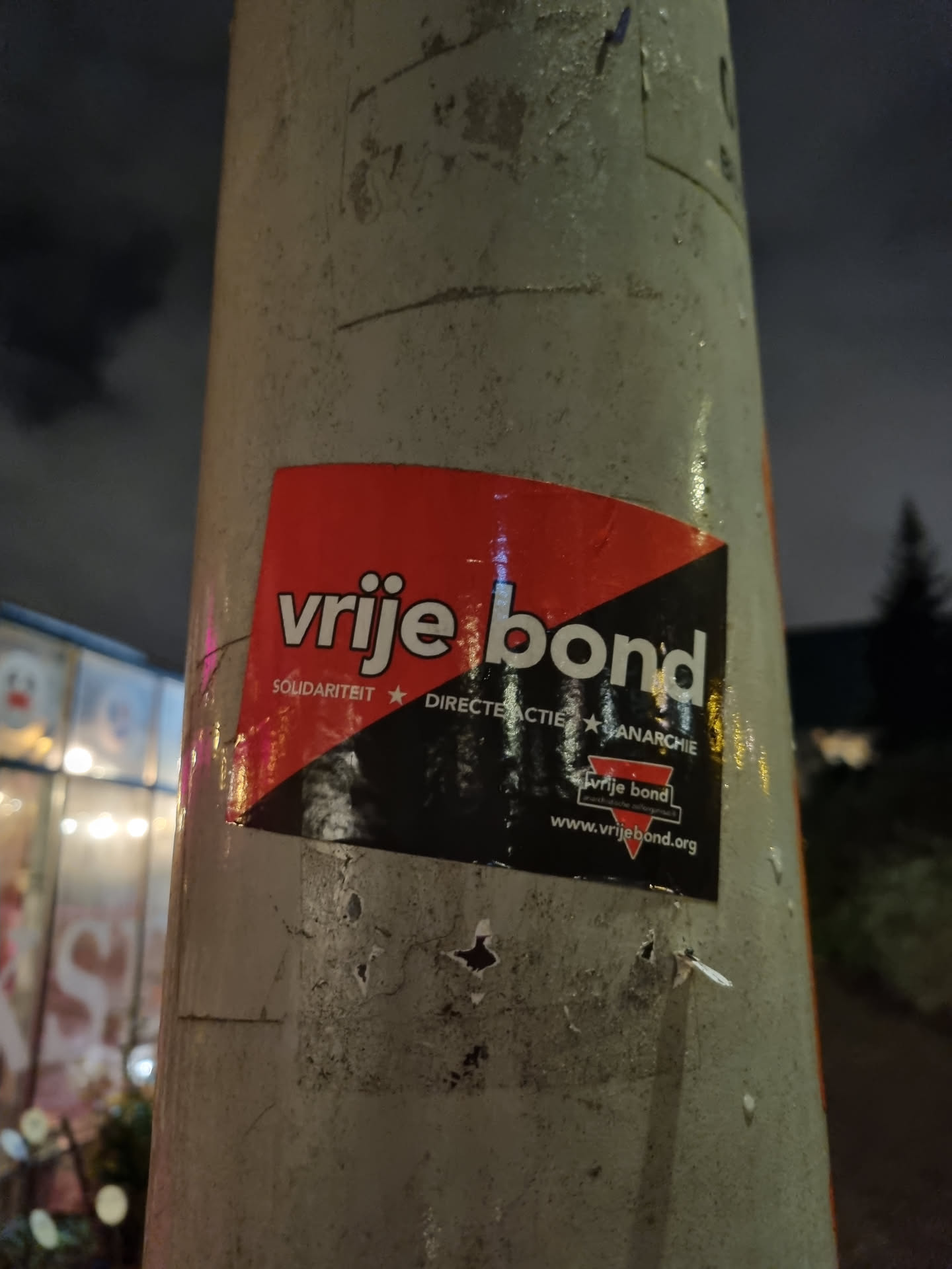 A sticker for VRIJE BOND (Free Union) which promotes solidarity, direct action and anarchy. The sticker is attached to a pole at night.