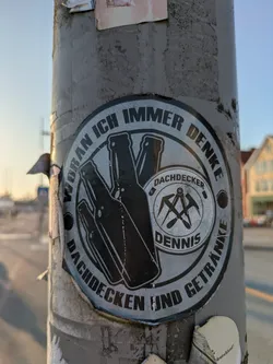 A weathered, circular black and white sticker is affixed to a grey metal pole. The sticker's design features a central inner circle containing the text 'DACHDECKER DENNIS' and a graphic of crossed roofing tools (likely a roofer's hammer and pick). Surrounding this inner circle, the main part of the sticker shows silhouettes of three beer bottles on the left, and a circular text that reads 'WORAN ICH IMMER DENKE' at the top and 'DACHDECKEN UND GETRÄNKE' at the bottom. The sticker appears to be peeling slightly at the edges.