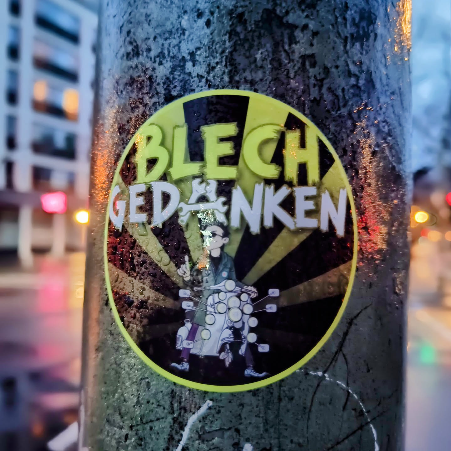 A round sticker with a cartoon drawing and the text "BLECH GEDANKEN". The sticker is affixed to a wet pole, and the background is blurred.