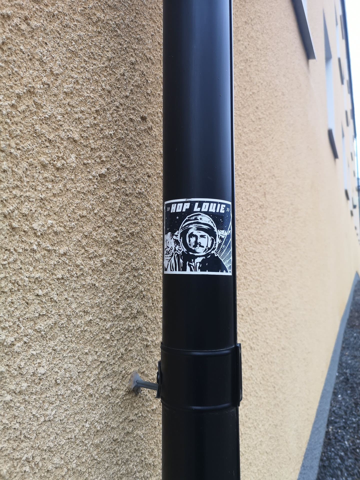 A rectangular, high-contrast black and white sticker featuring a stylized depiction of an astronaut or cosmonaut. The central figure wears a space helmet, revealing a face with a prominent mustache and a determined expression. The background is a dark, starry sky with abstract white lines or rays emanating from the right side. At the top, a black banner contains the white text "» HOP LOUIE »". The overall aesthetic is graphic and bold, reminiscent of retro sci-fi or propaganda art.