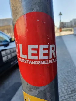 A red circular sticker affixed to a metal pole. The sticker features the word "LEER" (German for empty) in bold white letters, along with the website address "leerstandsmelder.de".  The website likely relates to reporting vacant properties or spaces in Germany. 