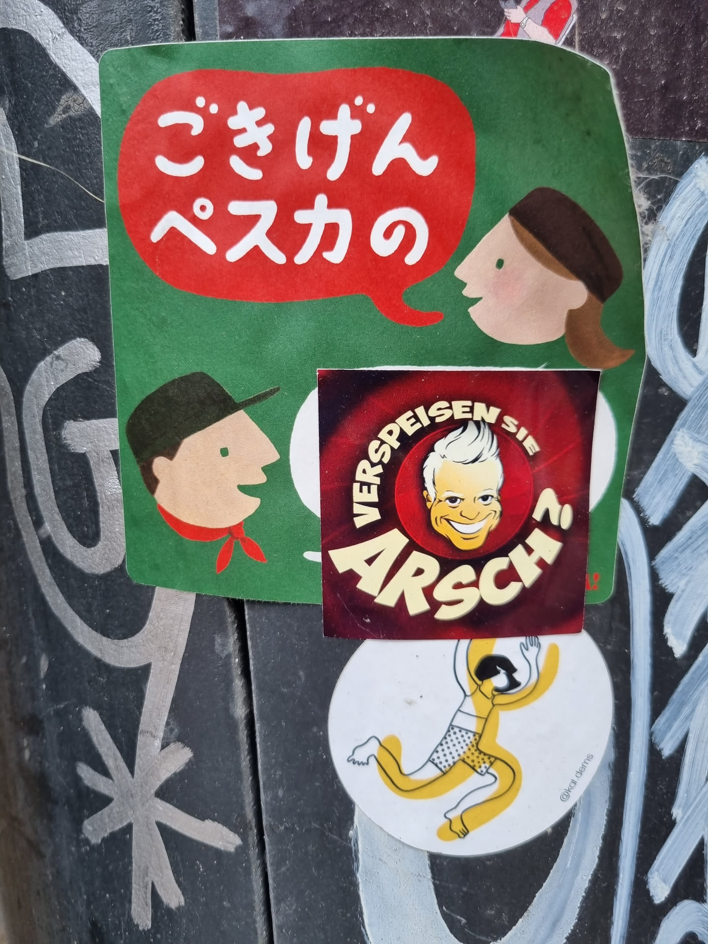 A collection of stickers on a dark surface. The main sticker features a speech bubble in red with Japanese text on a green background. There are two simple cartoon characters, one speaking in Japanese.  A smaller sticker underneath displays a cartoon man with blond hair, the word "Arsch" prominently displayed, and text in German around the edges.