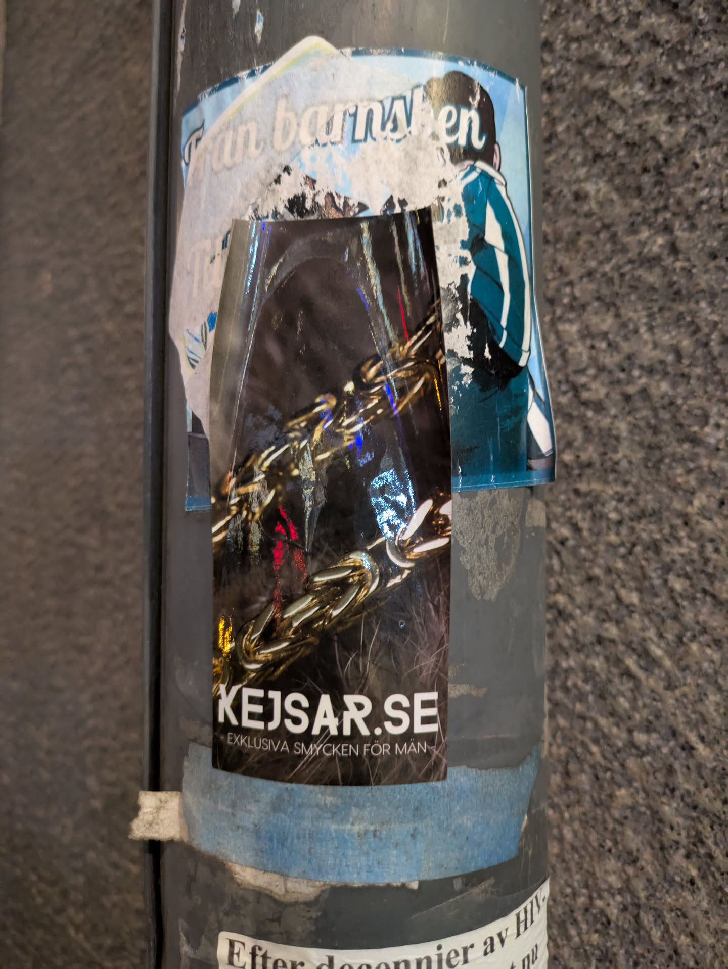 The sticker promotes KEJSAR.SE and their exclusive jewelry for men, showcasing a gold chain against a dark background. It is affixed to a pole with other stickers.