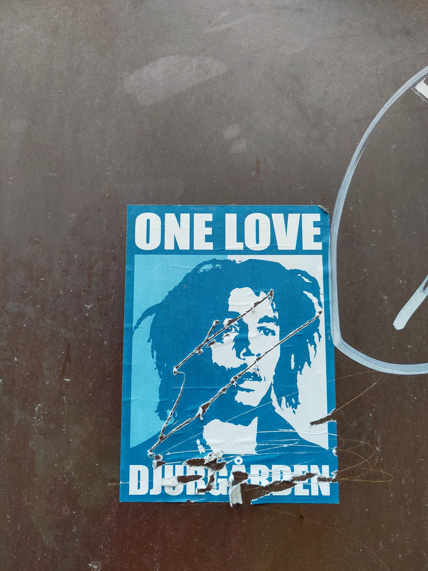 A sticker featuring Bob Marley's portrait with "One Love" and "Djurgården" written on it. The sticker shows some signs of wear and tear.