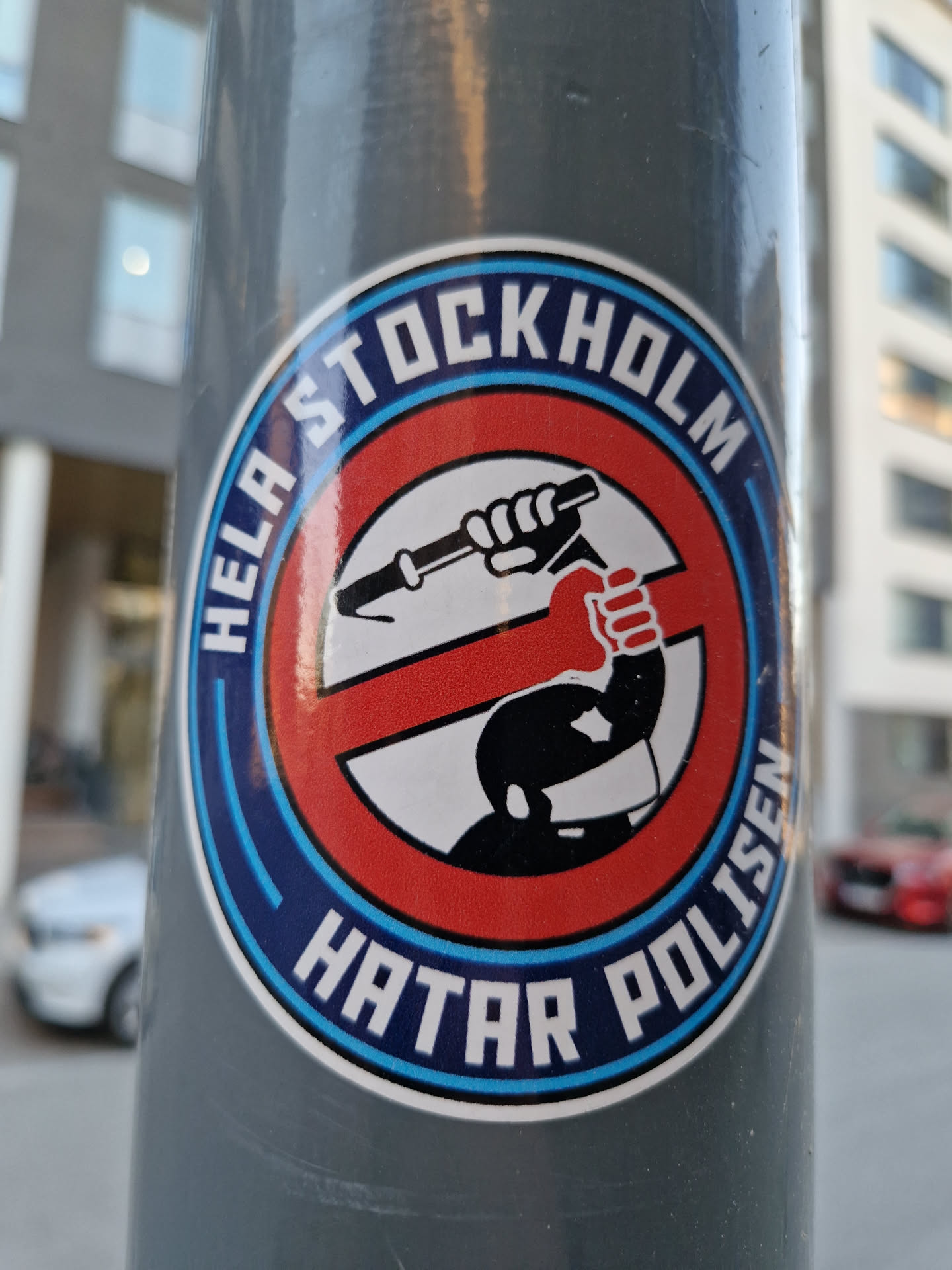 A round sticker with a red circle and a crossed-out image of a hand holding a baton. The text around the circle says "Hela Stockholm hatar polisen" which translates to "All of Stockholm hates the police".