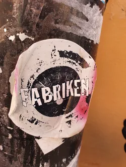 A worn sticker with the word "Fabriken" in large letters and a gauge image in the center. The sticker shows signs of wear and tear. It's attached to a dark-colored pole.