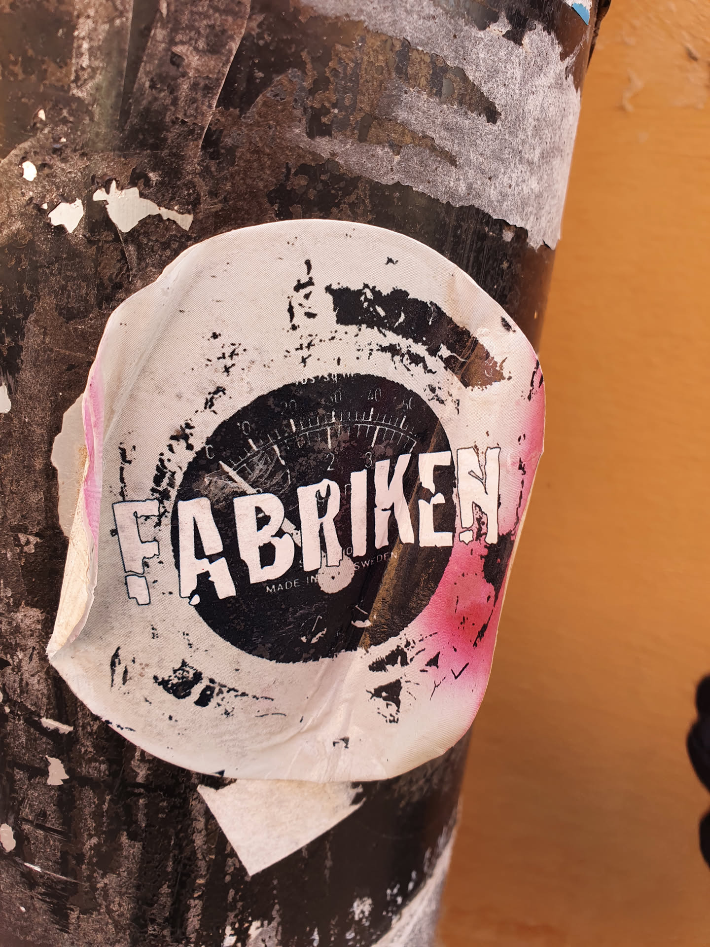 A worn sticker with the word "Fabriken" in large letters and a gauge image in the center. The sticker shows signs of wear and tear. It's attached to a dark-colored pole.