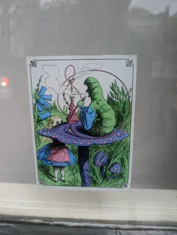 The sticker depicts a scene from Alice in Wonderland. A large, green, caterpillar-like creature with a blue robe sits on a purple mushroom with pink polka dots, smoking from a hookah. Smoke billows upwards, forming a circular pattern. To the left, a young girl, presumably Alice, with blonde hair and wearing a blue dress with a pink apron, peeks out from behind the mushroom. The background is filled with vibrant green grass and blue bell-shaped flowers. The overall style is illustrative and reminiscent of classic fantasy art, with bold colors and linework.