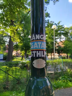 A sticker on a lamppost in Stockholm, Sweden. The sticker features the text "LÅNGA GATAM STHLM"  in blue and yellow, possibly referring to a street name.  The image shows lush green surroundings.