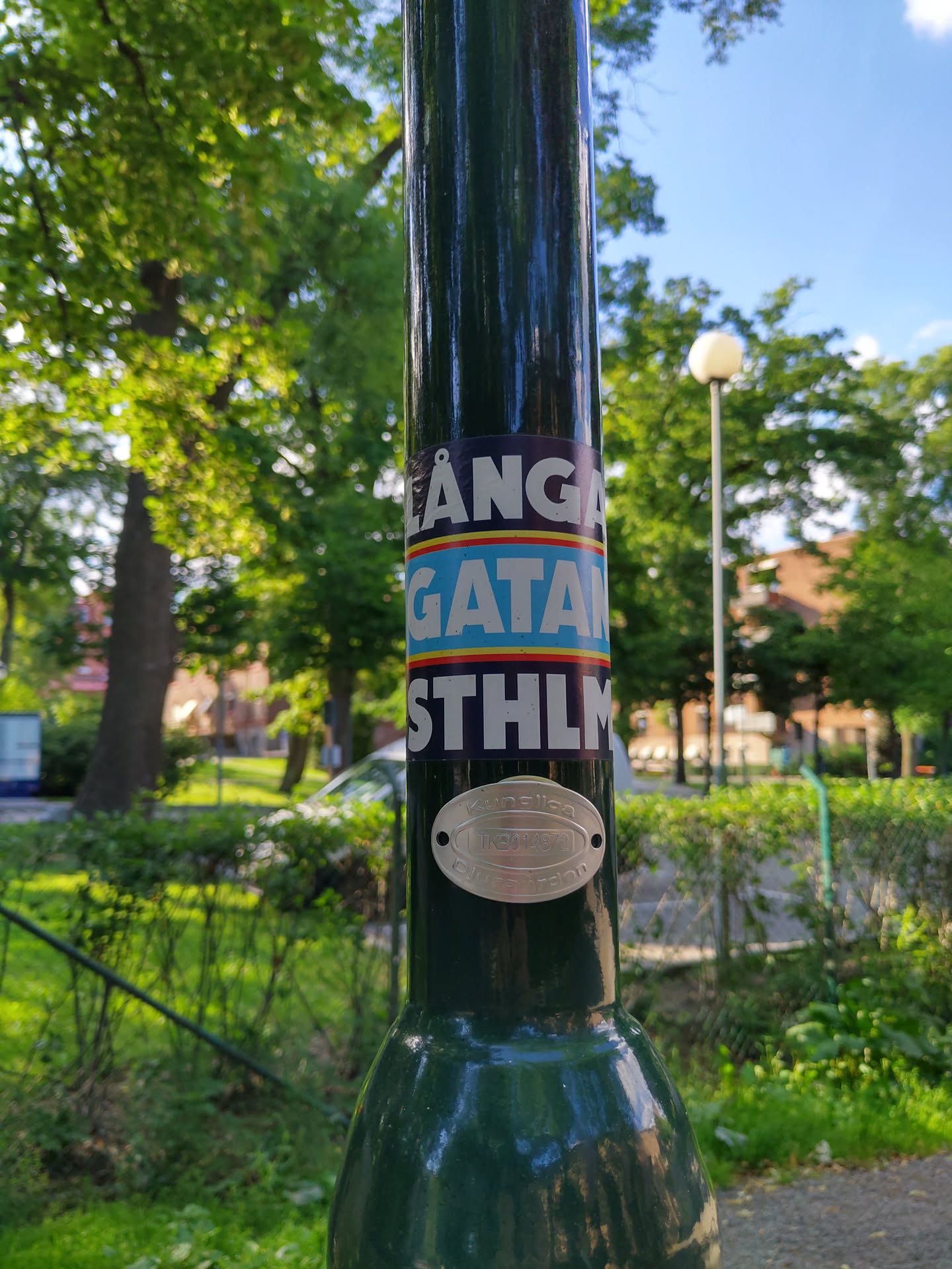 A sticker on a lamppost in Stockholm, Sweden. The sticker features the text "LÅNGA GATAM STHLM"  in blue and yellow, possibly referring to a street name.  The image shows lush green surroundings.