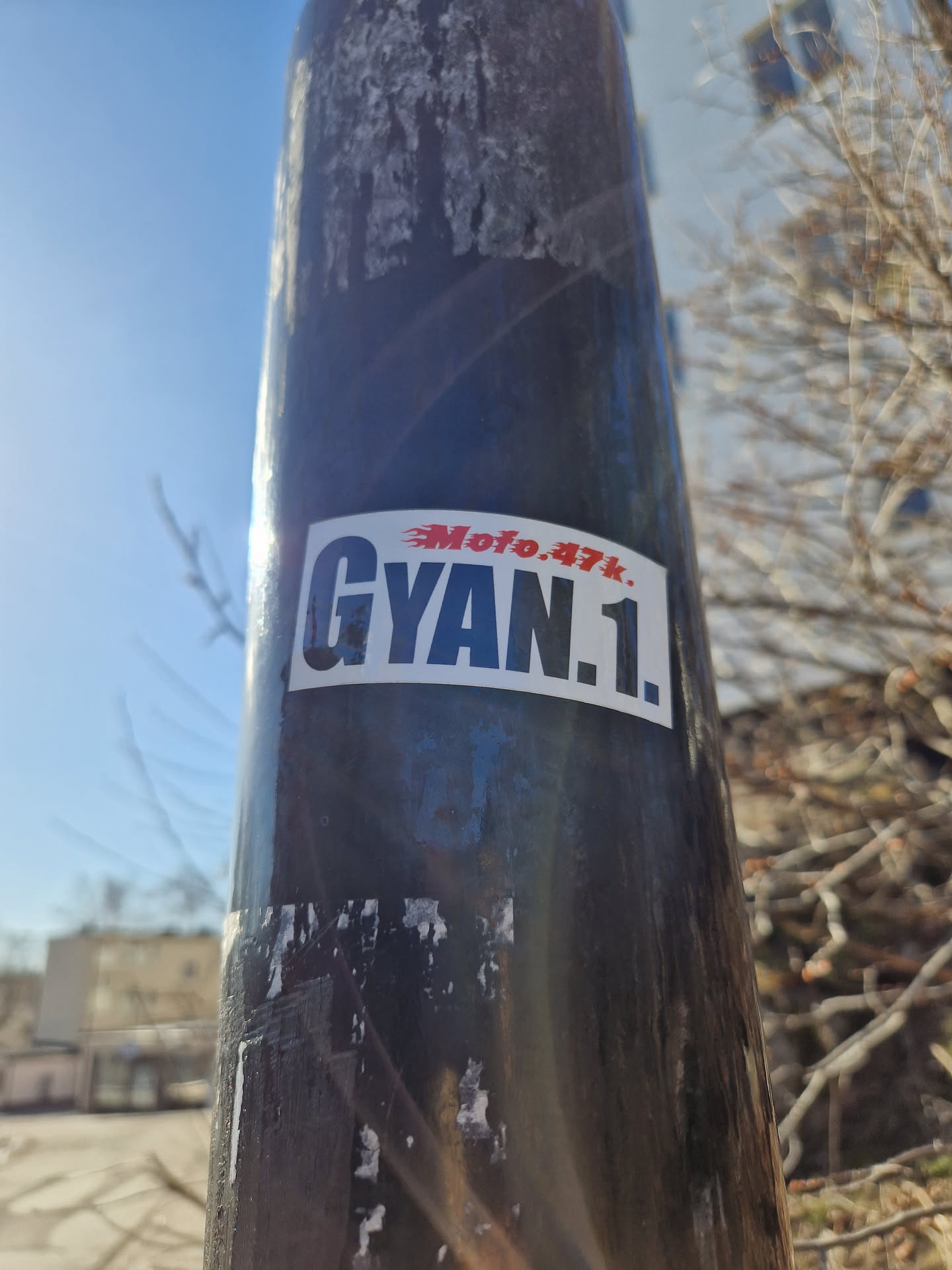 A sticker featuring the text \"GYAN.1\" with a smaller text \"Moto.47k\" above it. The sticker is placed on a dark pole outdoors.