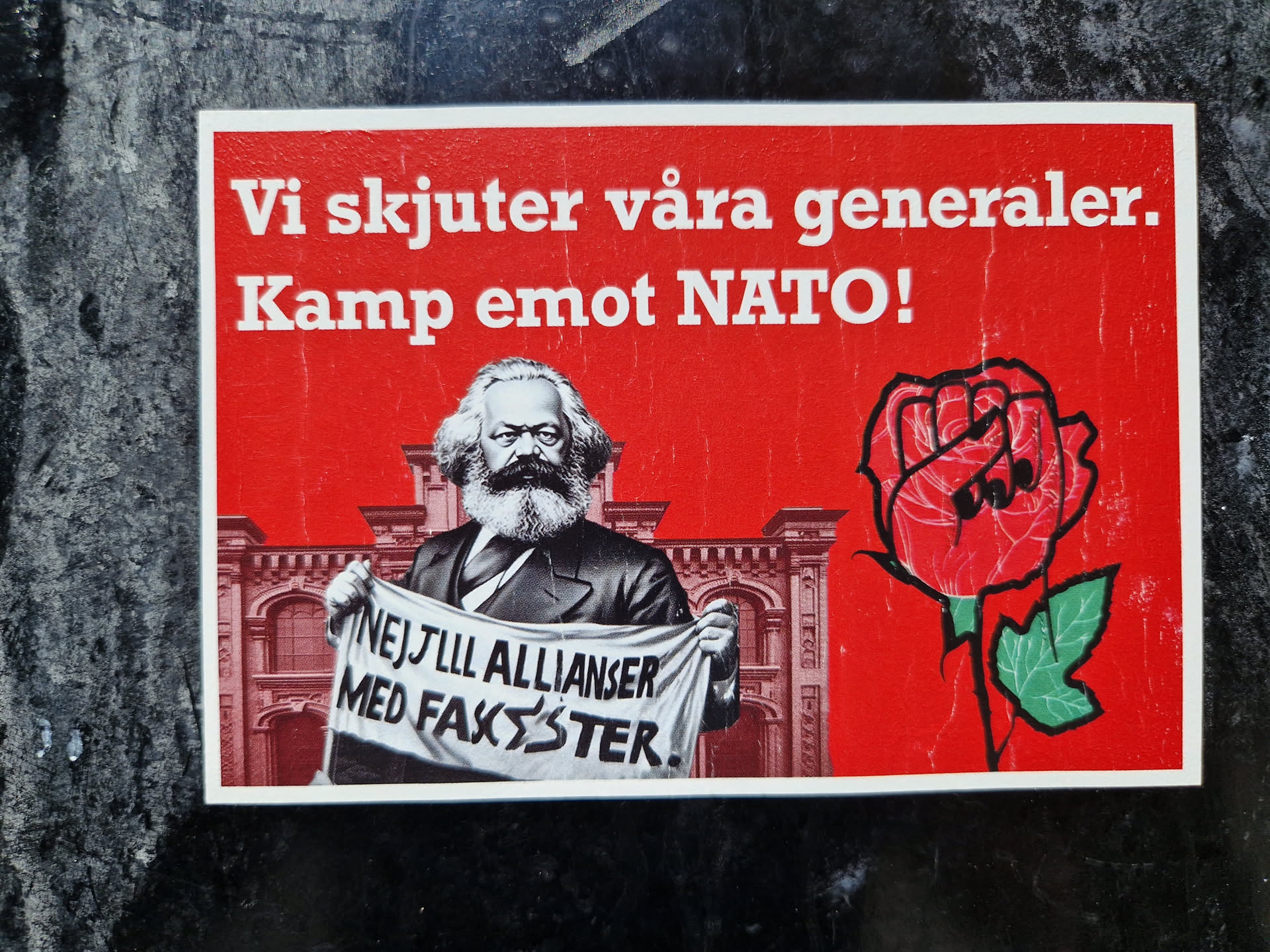 A sticker featuring Karl Marx holding a banner that says "Nej till allianser med fascister."  There's also a red rose with a fist in the middle, symbolizing resistance. The text on the sticker translates to "We shoot our generals. Fight against NATO!"