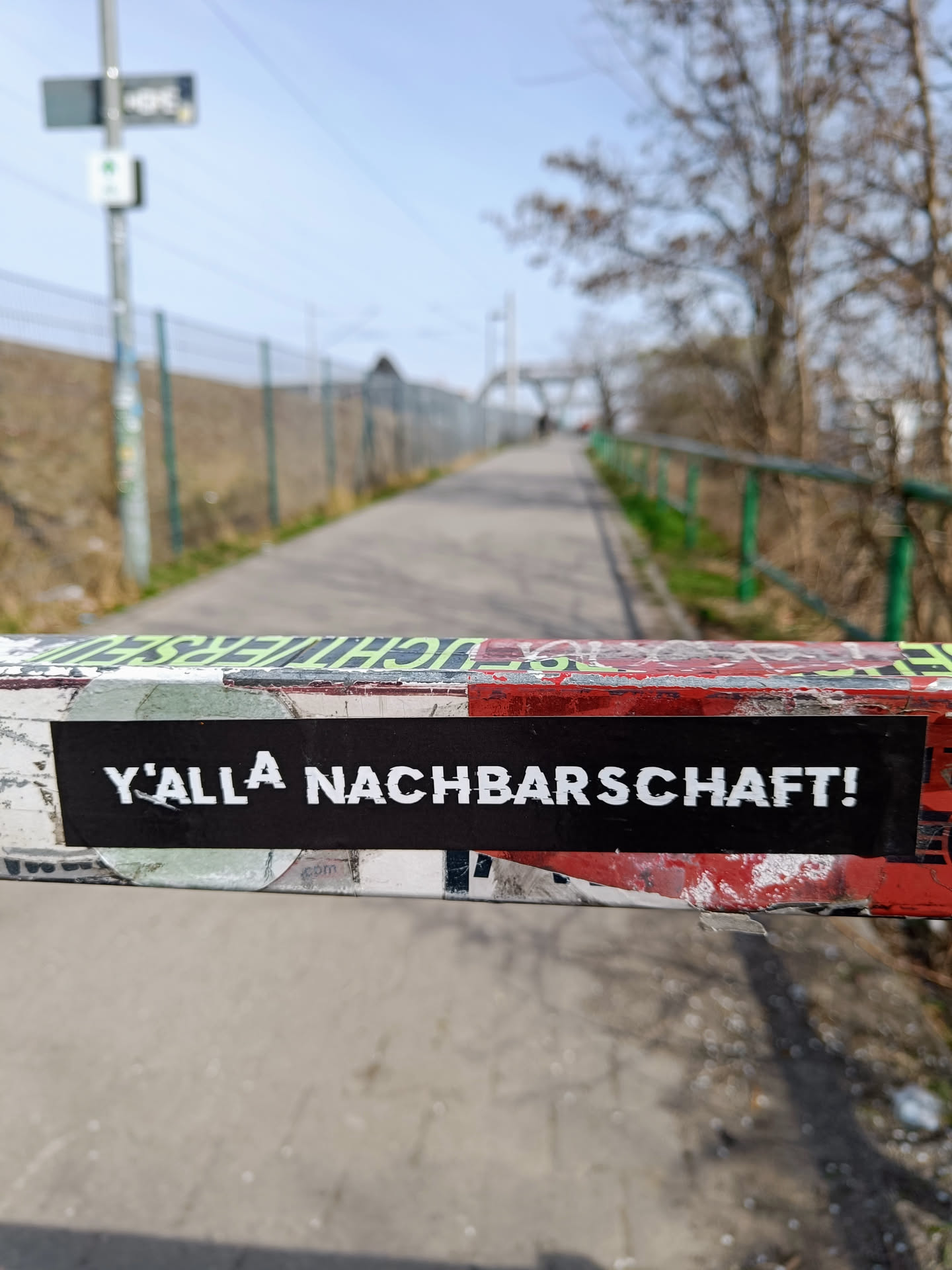 A black sticker with white lettering reading "Y'allA Nachbarschaft!"  affixed to a metal post. The sticker is slightly weathered, suggesting some time outdoors. The background is blurred, showing a paved path and trees. 