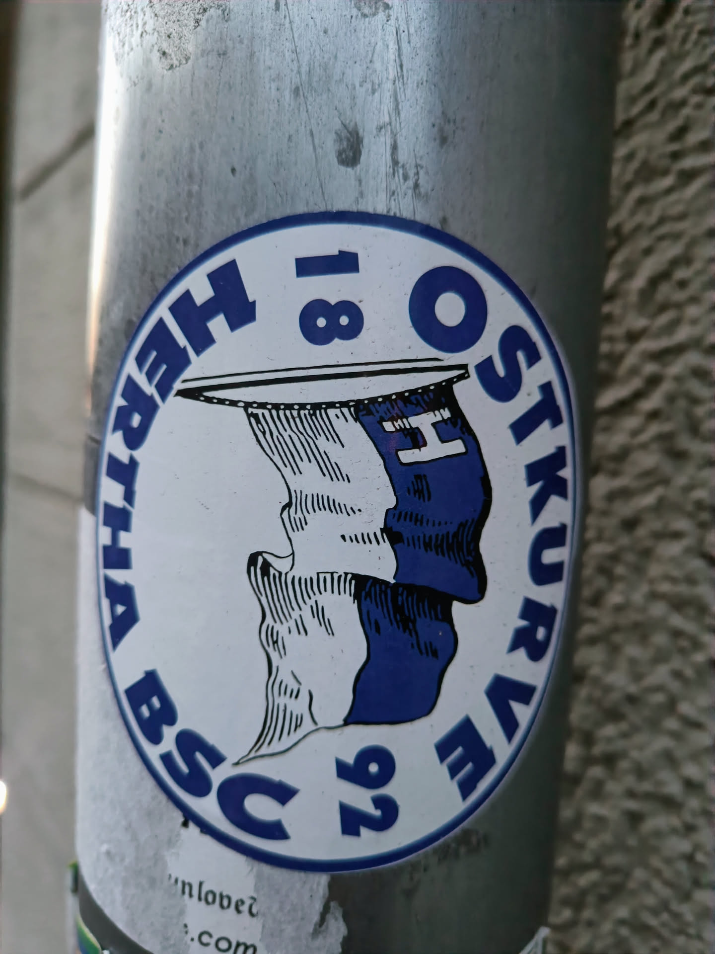 A round sticker featuring the logo of Hertha BSC's Ostkurve (East Stand) fan group.  The logo incorporates the club's name and founding year (1892), with a visual element that appears to depict a profile of a head. The colors are primarily white and blue.