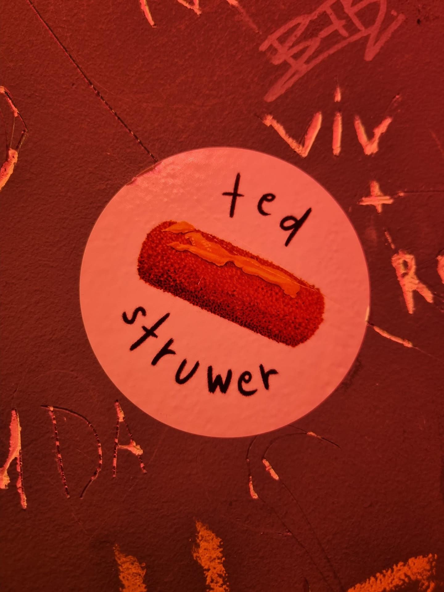 A round sticker with a drawing of a fried food, and the text "ted strüwer" written on it. The sticker is attached to a dark red wall with other graffiti around it.