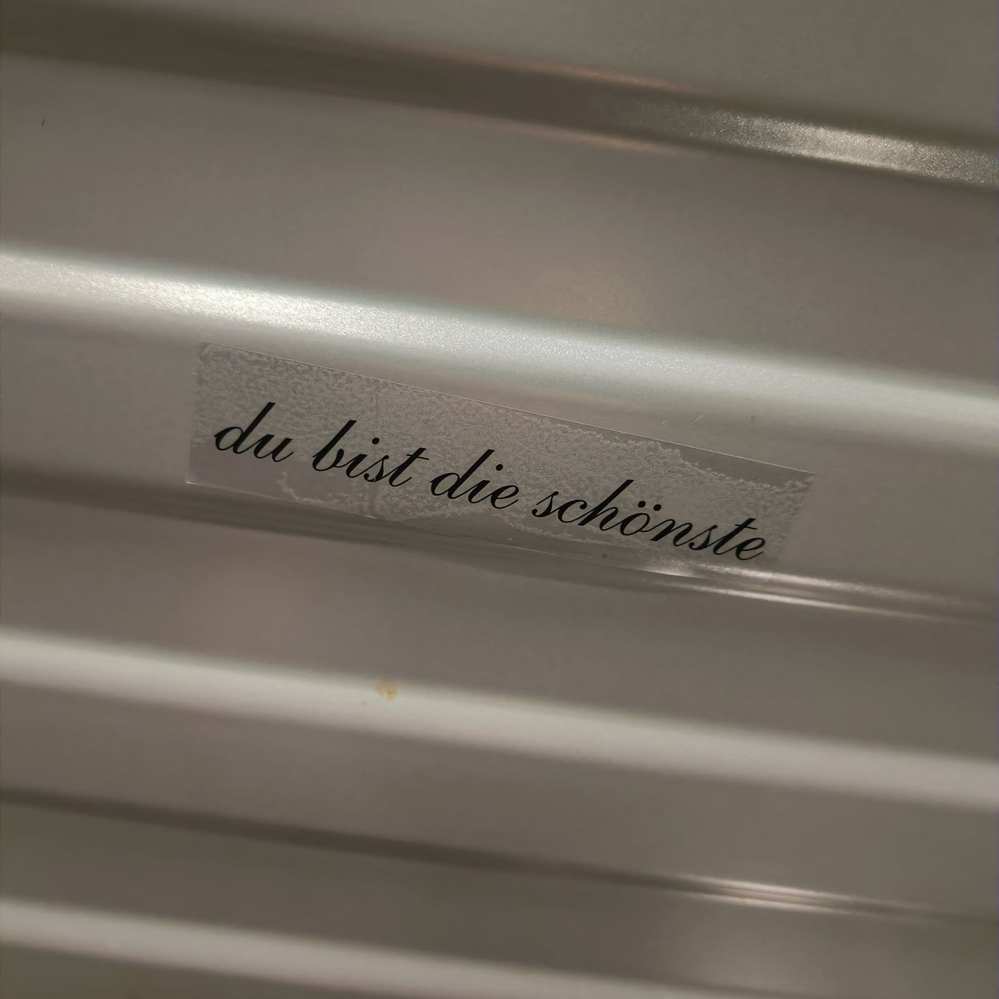 A small transparent sticker with the text "Du bist die schönste" (You are the most beautiful) written in a cursive font. The sticker is placed on a light gray ribbed surface.