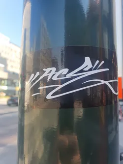 A black and white sticker featuring a stylized signature or logo. The lettering is white on a black background. The sticker is applied to a dark green pole. 