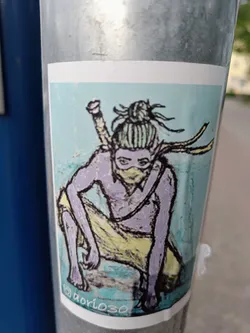 A sticker featuring an illustration of a ninja-like character crouching, holding a sword. The character has purple skin, yellow clothing, and a topknot. The art style is hand-drawn, with visible texture and color variations.
