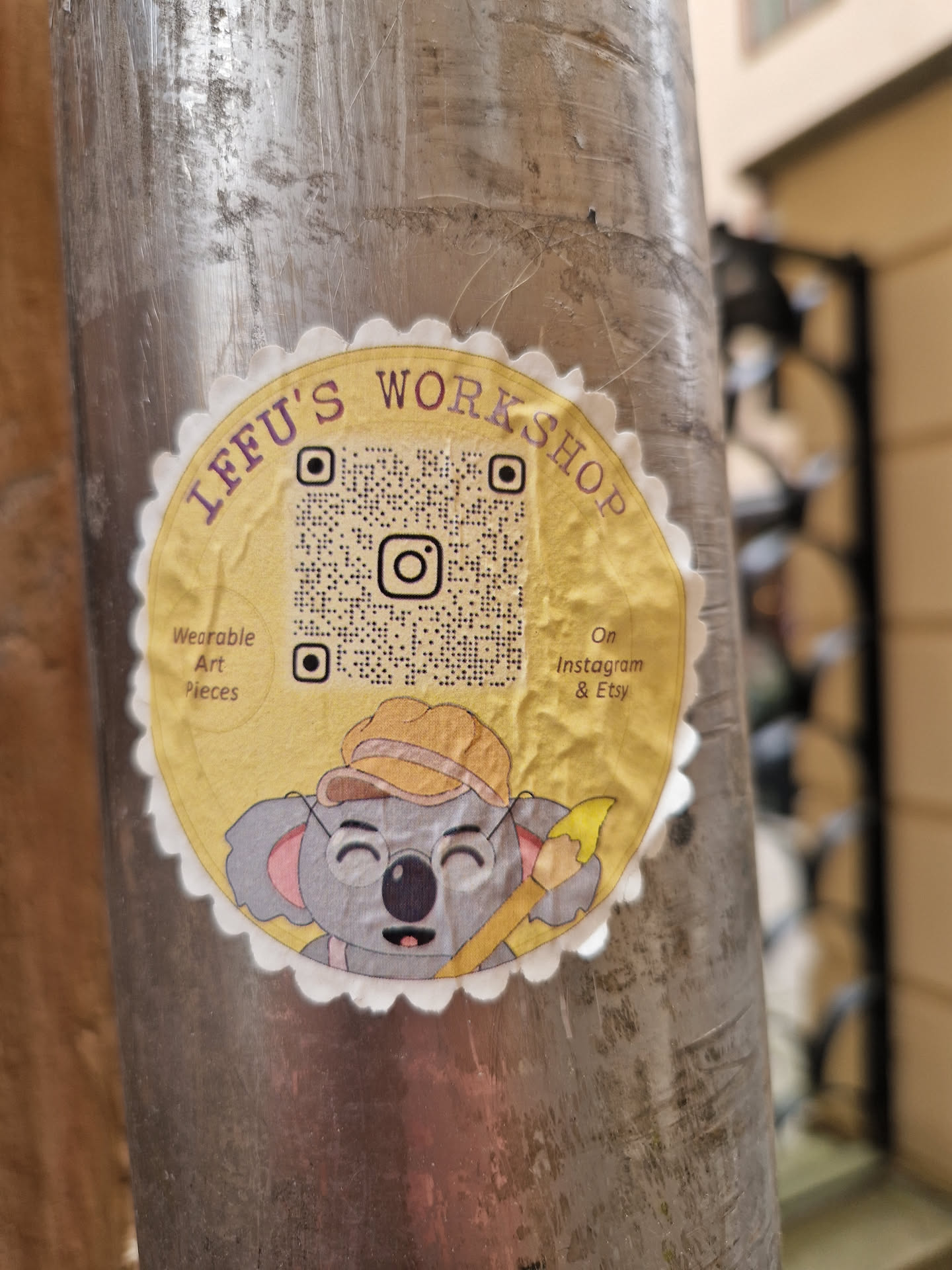 Round sticker featuring an illustration of a koala wearing a hat and holding a paintbrush. The sticker has a yellow background with text that says "LFFU's Workshop", along with a QR code and information about where to find their wearable art pieces (Instagram & Etsy).