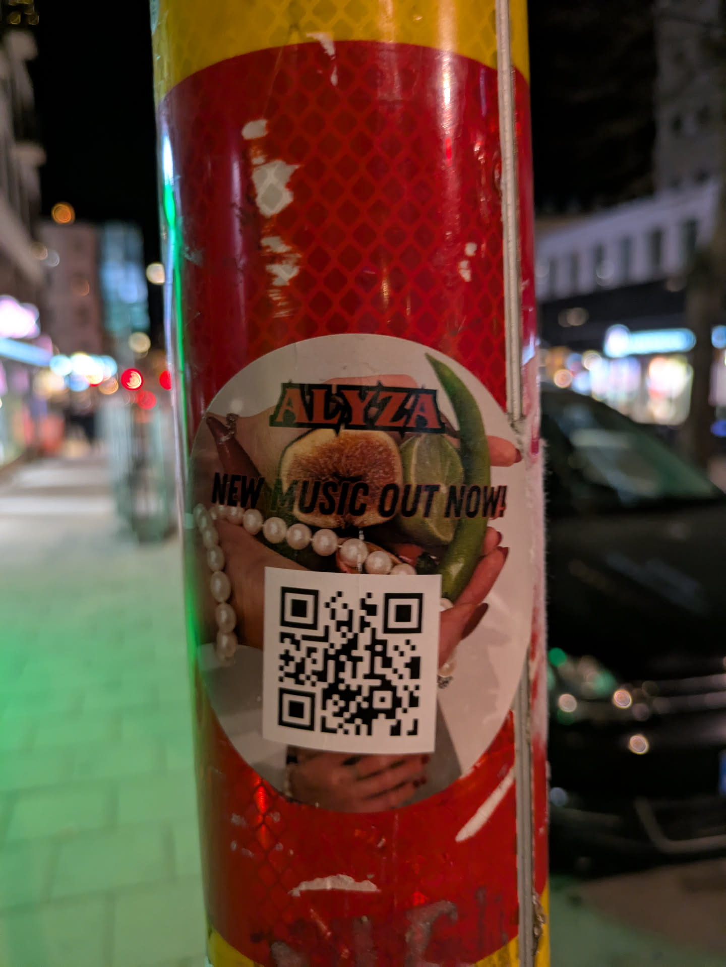 A round sticker is affixed to a pole with reflective red and yellow stripes. The sticker features the name "ALYZZA" in a stylized font above an image of hands holding a fig, a lime and a green chili. The text "NEW MUSIC OUT NOW!" is printed on the sticker. There is also a QR code present.