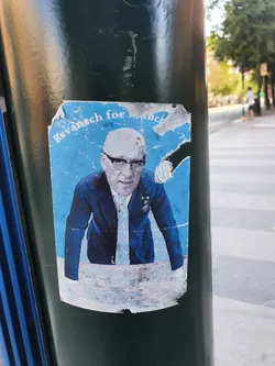 A sticker featuring a stylized image of a man in a suit, possibly referencing a historical event related to Munich. The sticker is somewhat damaged and affixed to a street pole.