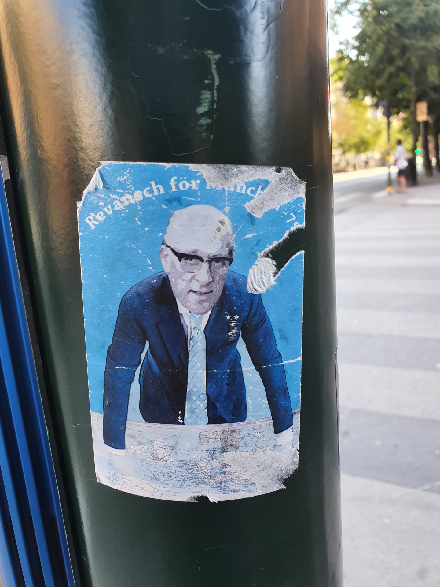 A sticker featuring a stylized image of a man in a suit, possibly referencing a historical event related to Munich. The sticker is somewhat damaged and affixed to a street pole.