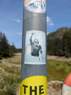 A faded and worn sticker affixed to a textured metal pole, featuring a stylized black and white depiction of a male figure, likely a football player, with long hair and his right arm raised in a celebratory gesture. The figure is wearing a jersey with text that appears to be "misuralla". Below the image, there is a clear white banner with black text that reads "ROMA CLUB NORVEGIA". The sticker shows signs of age and wear around its edges.
