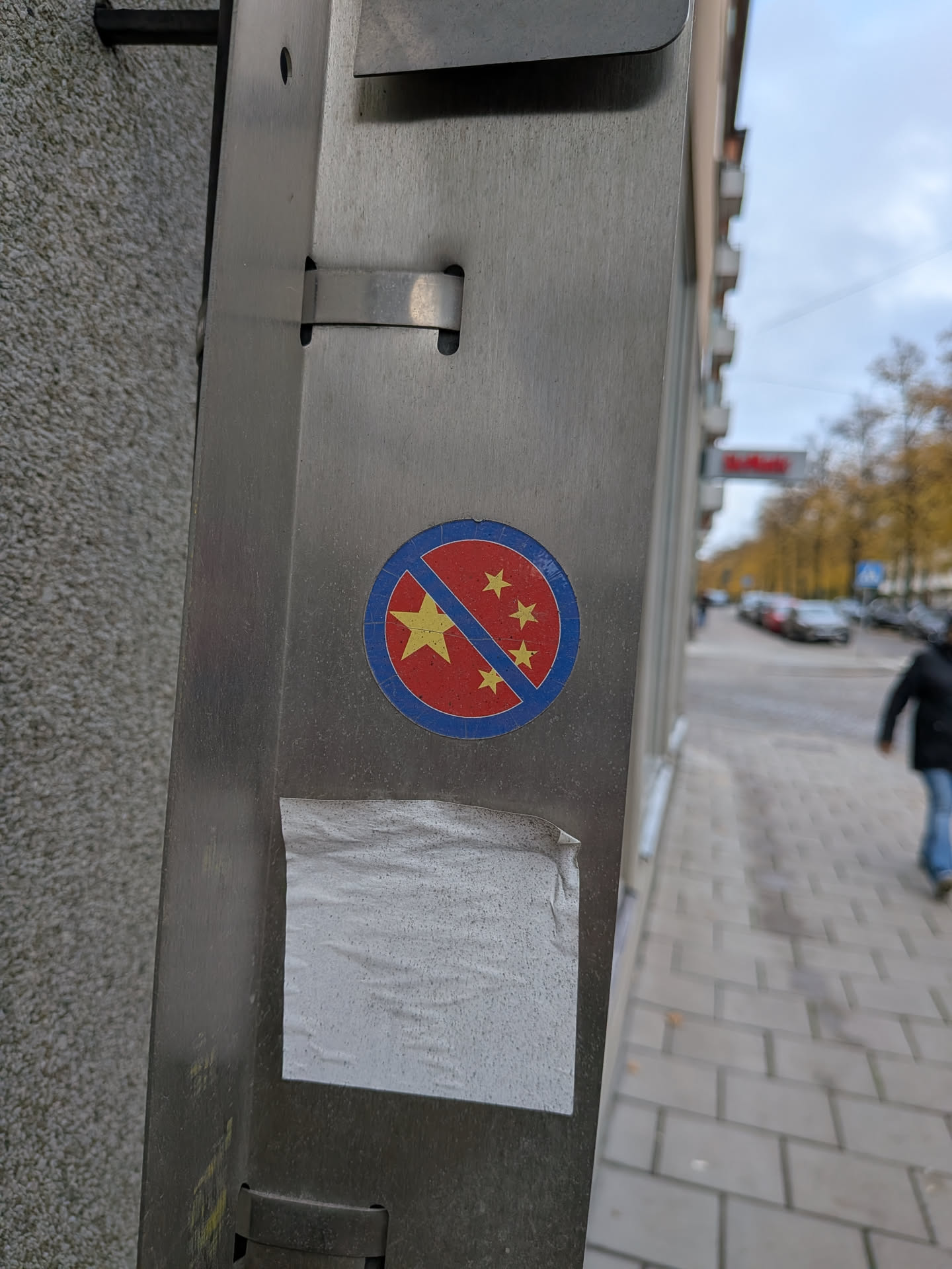 A circular sticker affixed to a brushed metal post. The sticker features a red center framed by a blue circular border with a diagonal blue line crossing across it from top right to bottom left, mimicking a prohibition symbol. Inside the red field are five yellow five-pointed stars arranged similarly to the flag of China—one large star on the left and four smaller stars in an arc on the right.