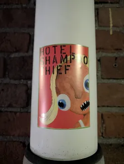 Street sticker This sticker is on a white pole and features a cartoon drawing of a character with exaggerated eyes, sharp teeth, and blonde hair, set against a red background. Text above the image reads "HOTEL SHAMPOO THIEF".