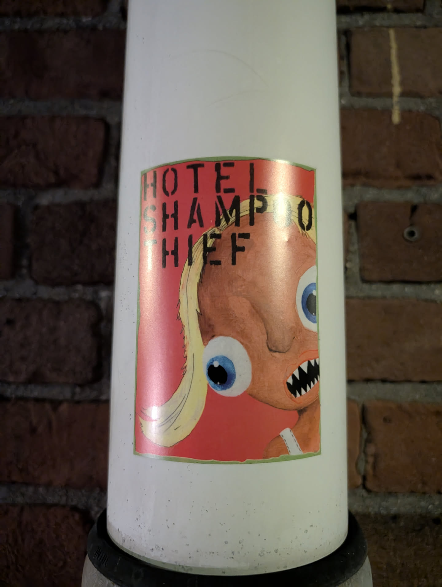 This sticker is on a white pole and features a cartoon drawing of a character with exaggerated eyes, sharp teeth, and blonde hair, set against a red background. Text above the image reads "HOTEL SHAMPOO THIEF".