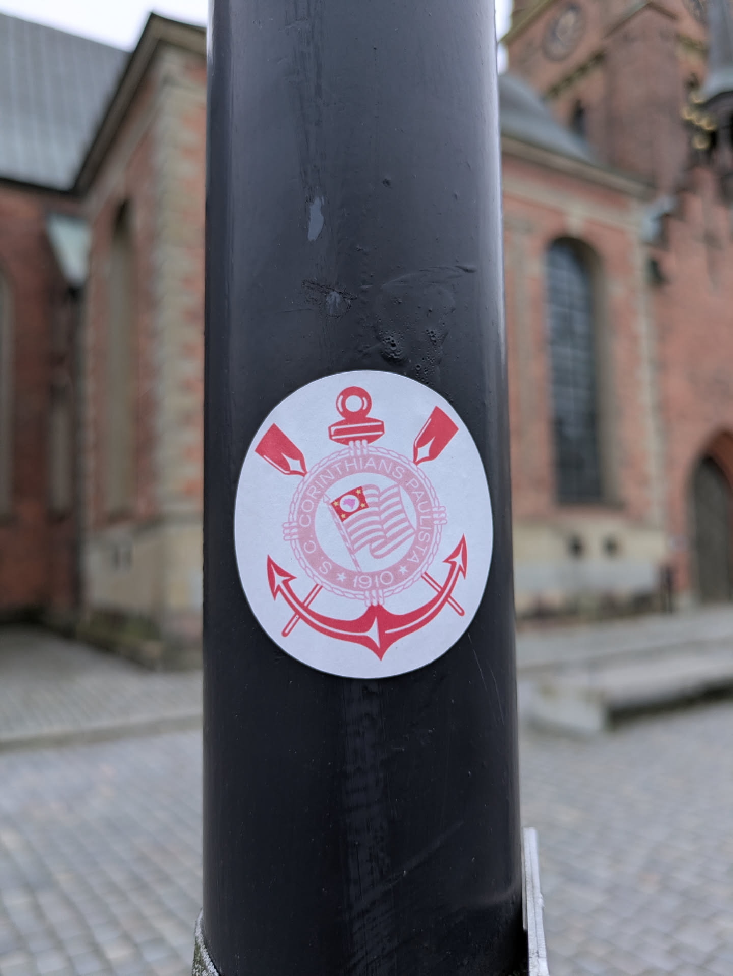 A round sticker featuring the logo of Sport Club Corinthians Paulista, a Brazilian football club. The logo is primarily red and white, and includes an anchor and crossed oars. The sticker is affixed to a dark-colored lamppost outside.  The background shows an out of focus brick building.