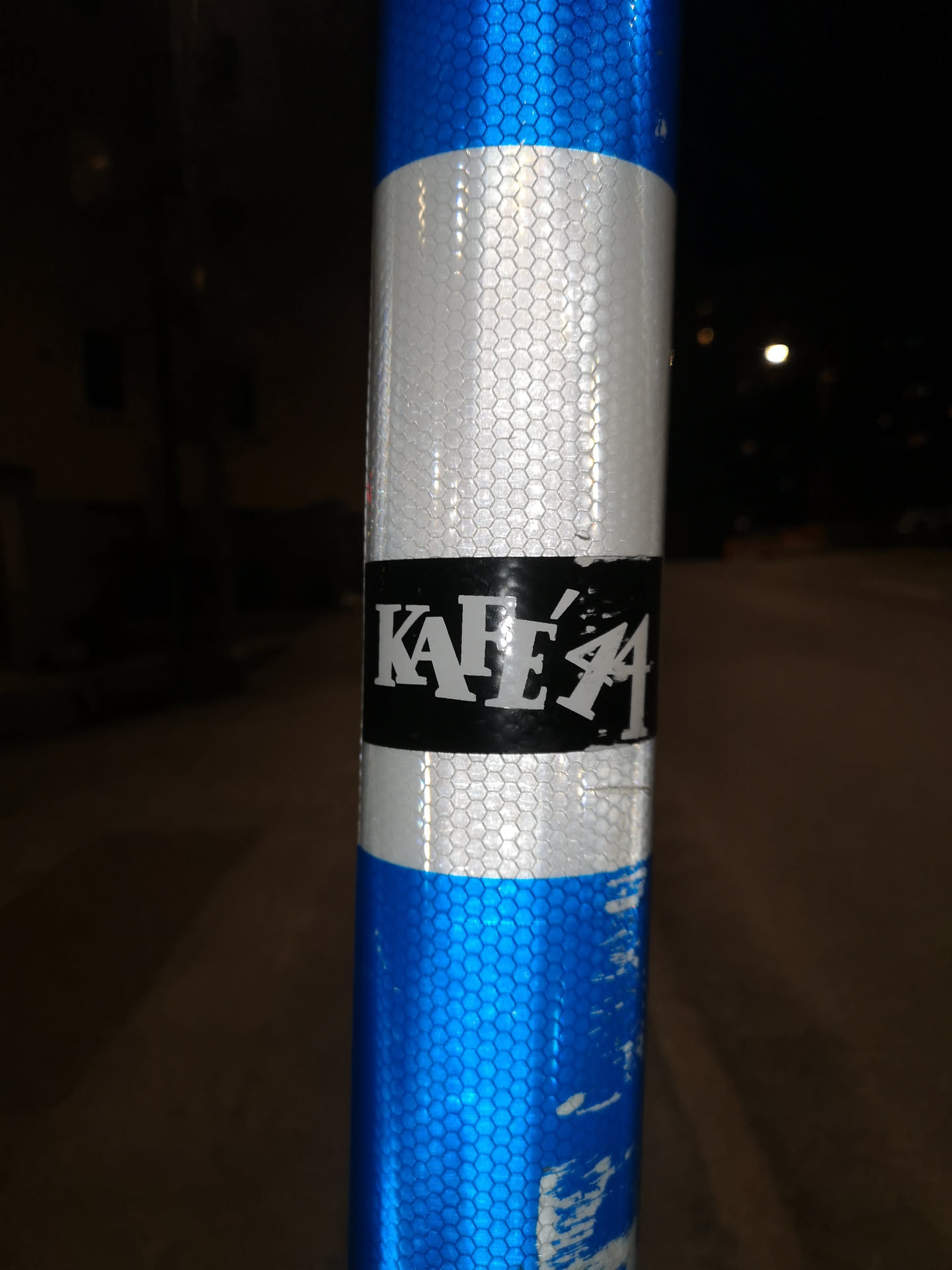 The sticker is rectangular with a solid black background. It features prominent white, uppercase, sans-serif text that reads "KAFÉ'44". The 'E' has an acute accent, and the '44' is stylized, appearing slightly integrated or overlapping with the 'E'. The sticker is applied to a silver reflective section of a pole, which has a honeycomb pattern. The edges of the sticker show some minor wear and tear.