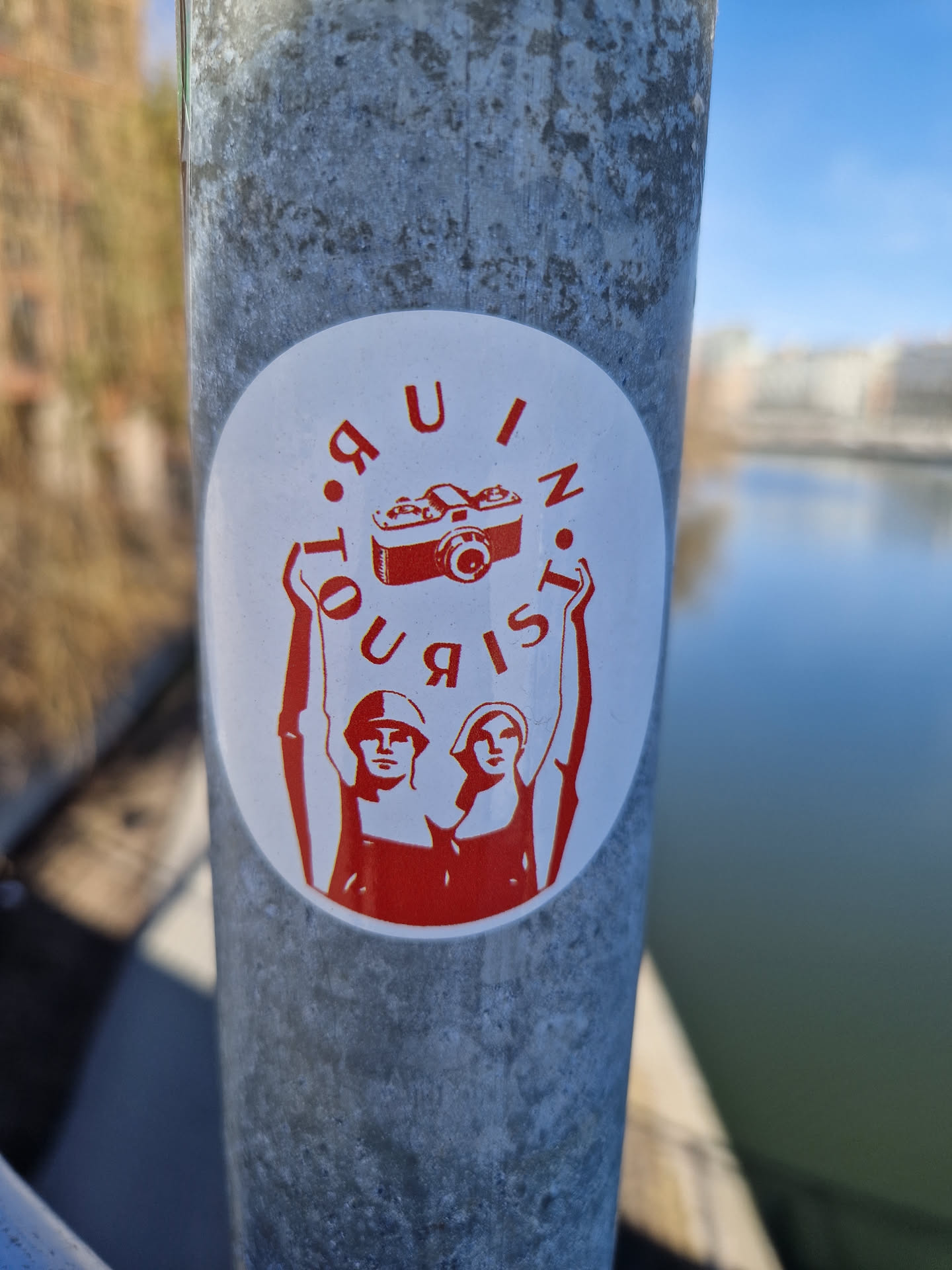 A red and white round sticker with an image of two people raising their arms, a camera, and the words "Tourist in Ruin" written in a circle around the image.