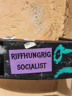 A purple sticker with black text on it. The text says "Riffhungrig Socialist". The sticker is attached to a black metal surface.