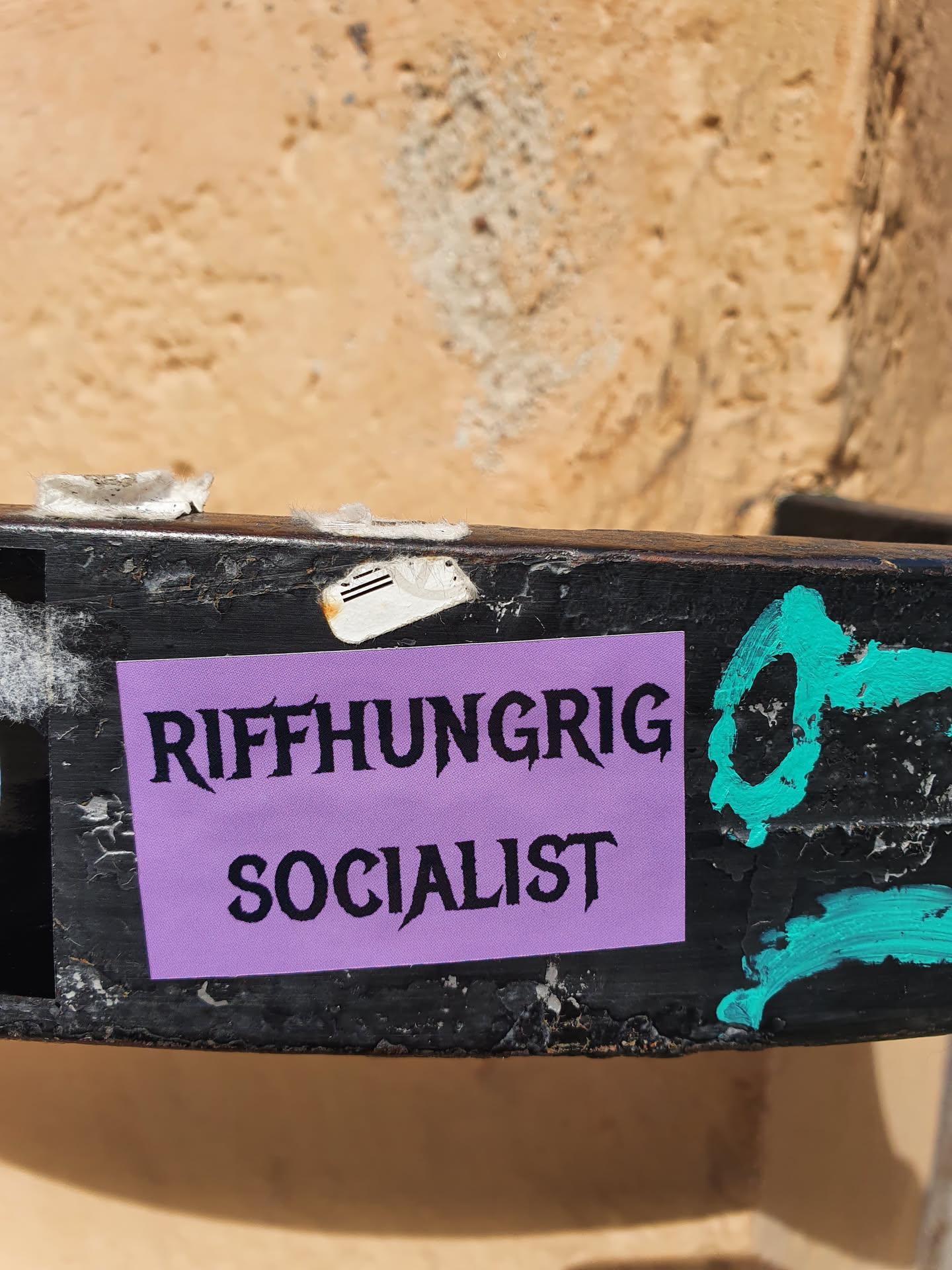 A purple sticker with black text on it. The text says "Riffhungrig Socialist". The sticker is attached to a black metal surface.