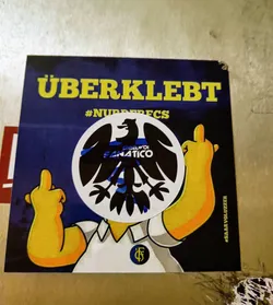 Sticker featuring a Simpsons character with a black eagle emblem replacing its head. The eagle emblem has the text "Engelvoi Fanatico" written inside. The background is dark blue with yellow text that says "ÜBERKLEBT" and the hashtag  "#NURDERFCS".  The Simpsons character is giving the middle finger with both hands.