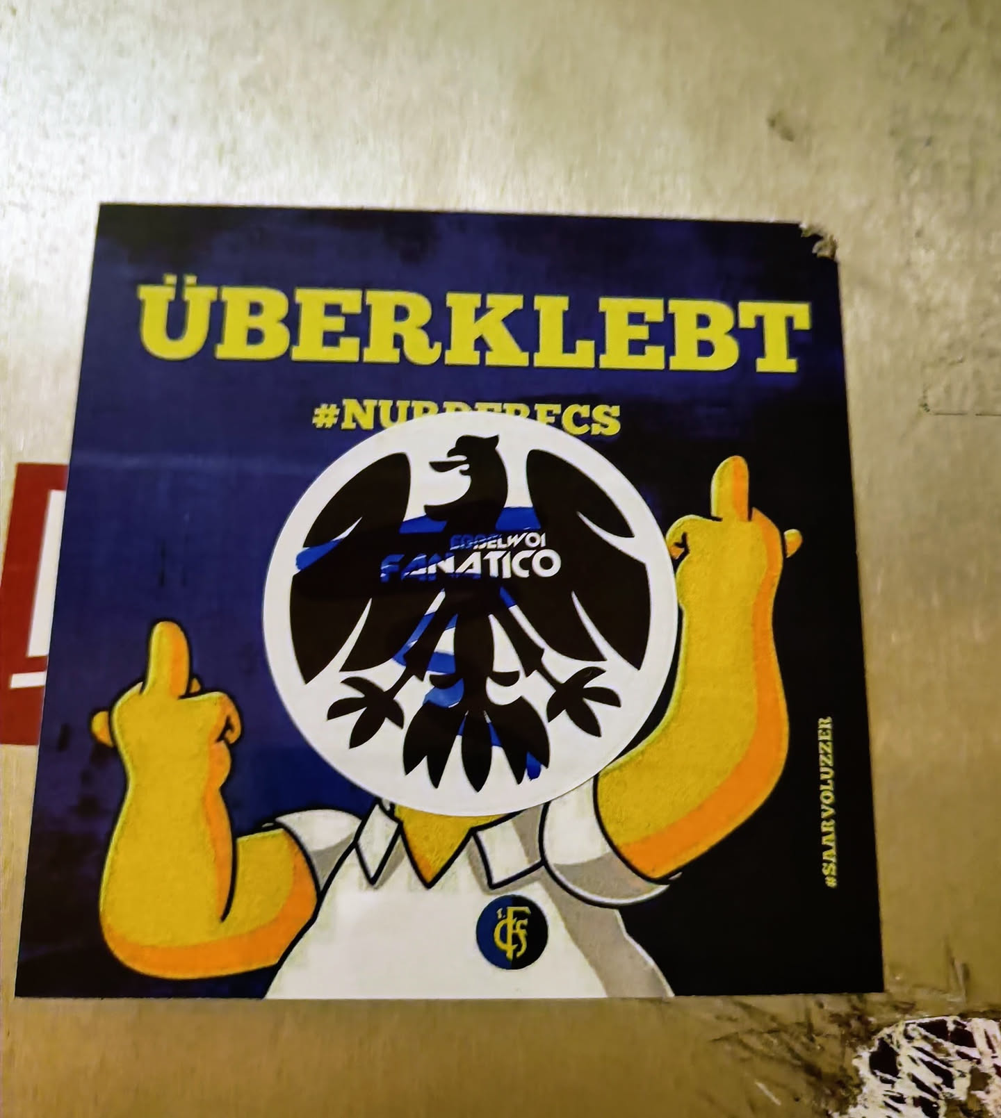 Sticker featuring a Simpsons character with a black eagle emblem replacing its head. The eagle emblem has the text "Engelvoi Fanatico" written inside. The background is dark blue with yellow text that says "ÜBERKLEBT" and the hashtag  "#NURDERFCS".  The Simpsons character is giving the middle finger with both hands.