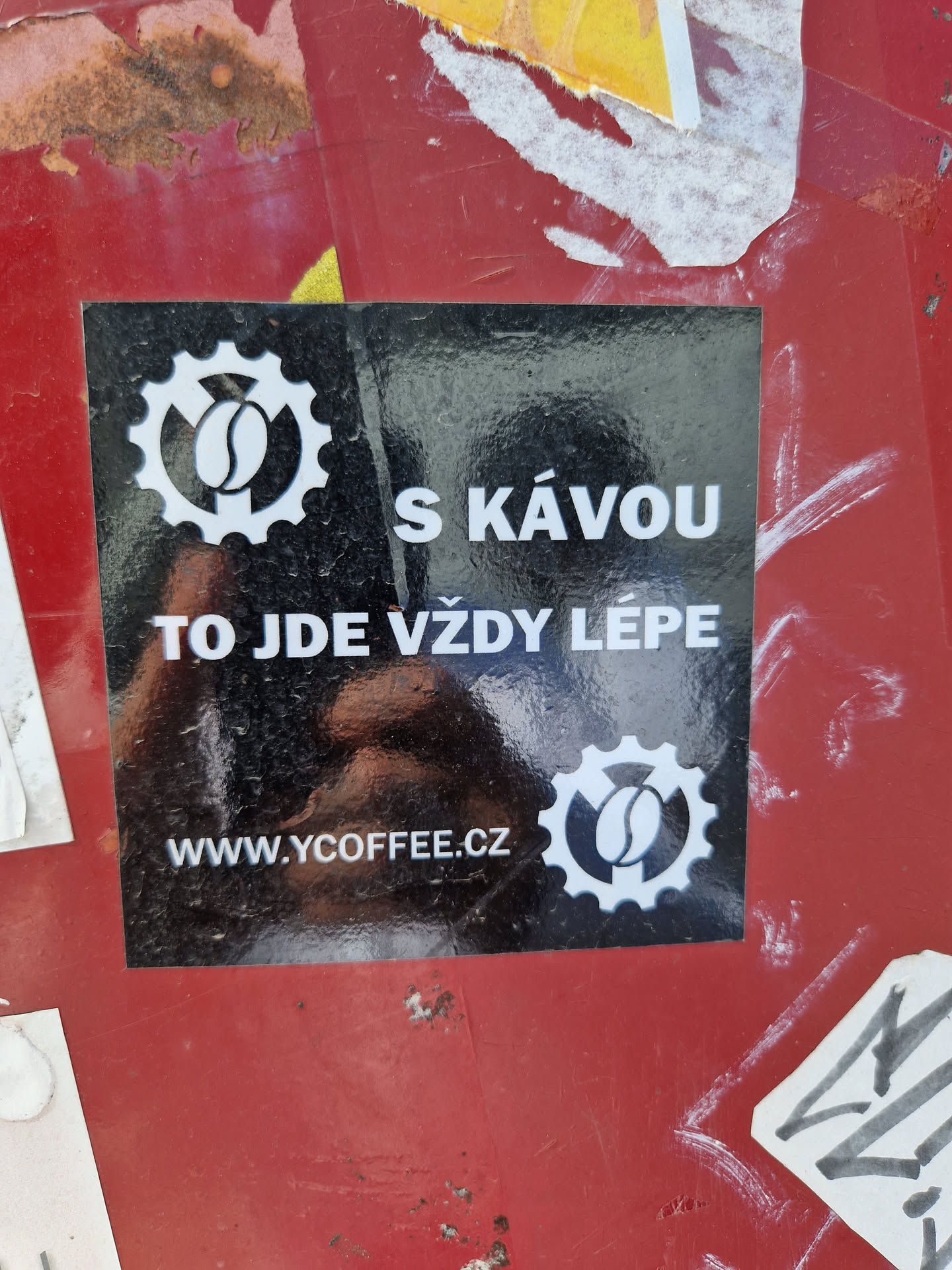Black sticker with white text and logo.  The logo features a coffee bean inside a gear. The text on the sticker says "S kávou to jde vždy lépe" which translates to "With coffee, it's always better" in English.  A website address is also included at the bottom.
