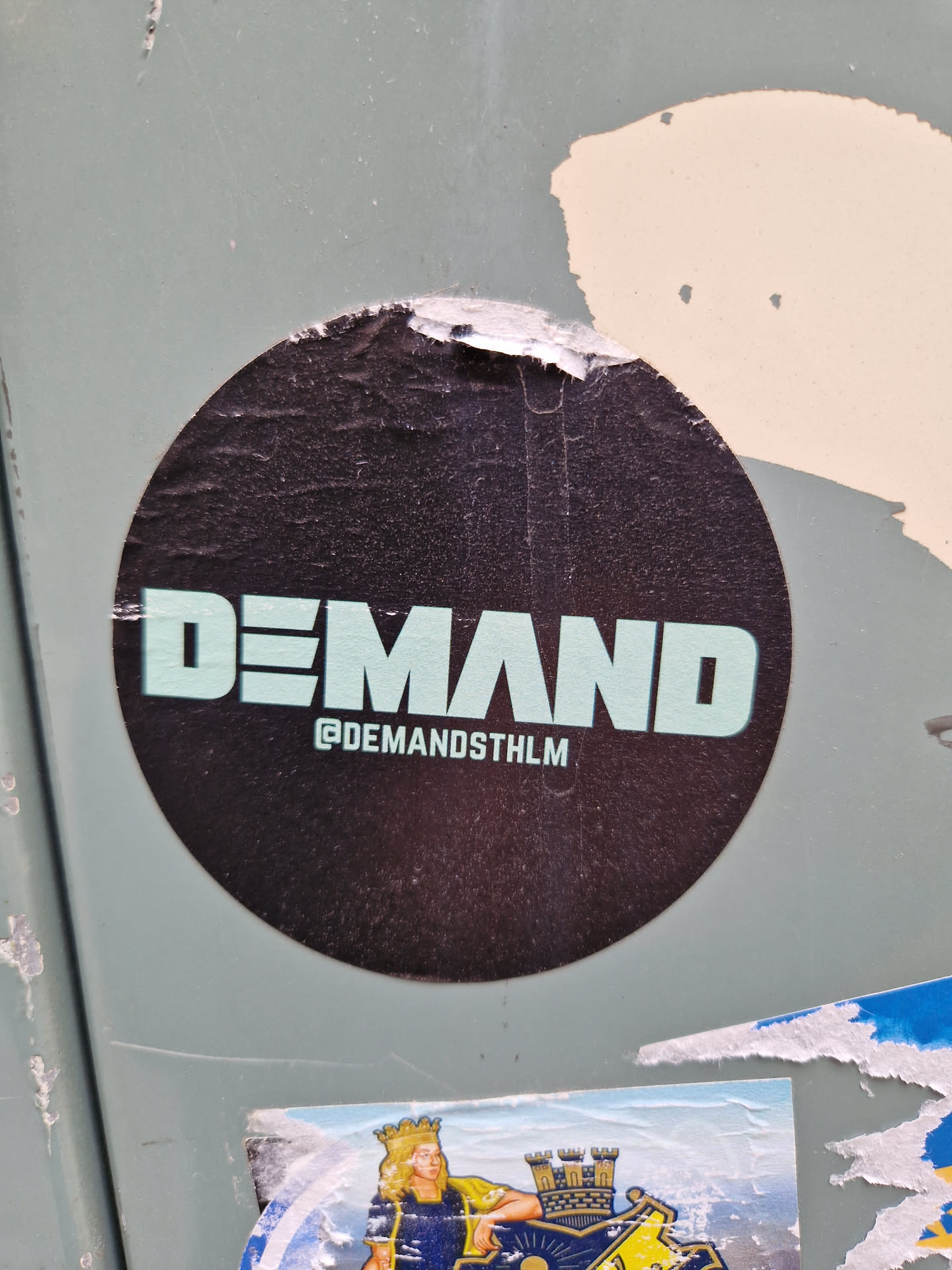 Round black sticker with the word DEMAND written in a teal sans-serif font. Below the text is the instagram handle @DEMANDSTHLM.