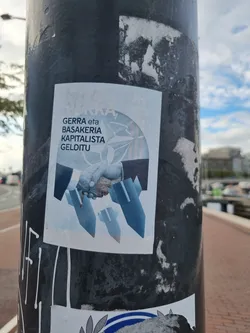 A sticker with a design that depicts a handshake between two figures, with what looks like weapons or tools emerging from beneath their clasped hands. The text on the sticker translates to "Stop War and Capitalist Barbarity". The style is minimalist, using shades of blue on a white background.