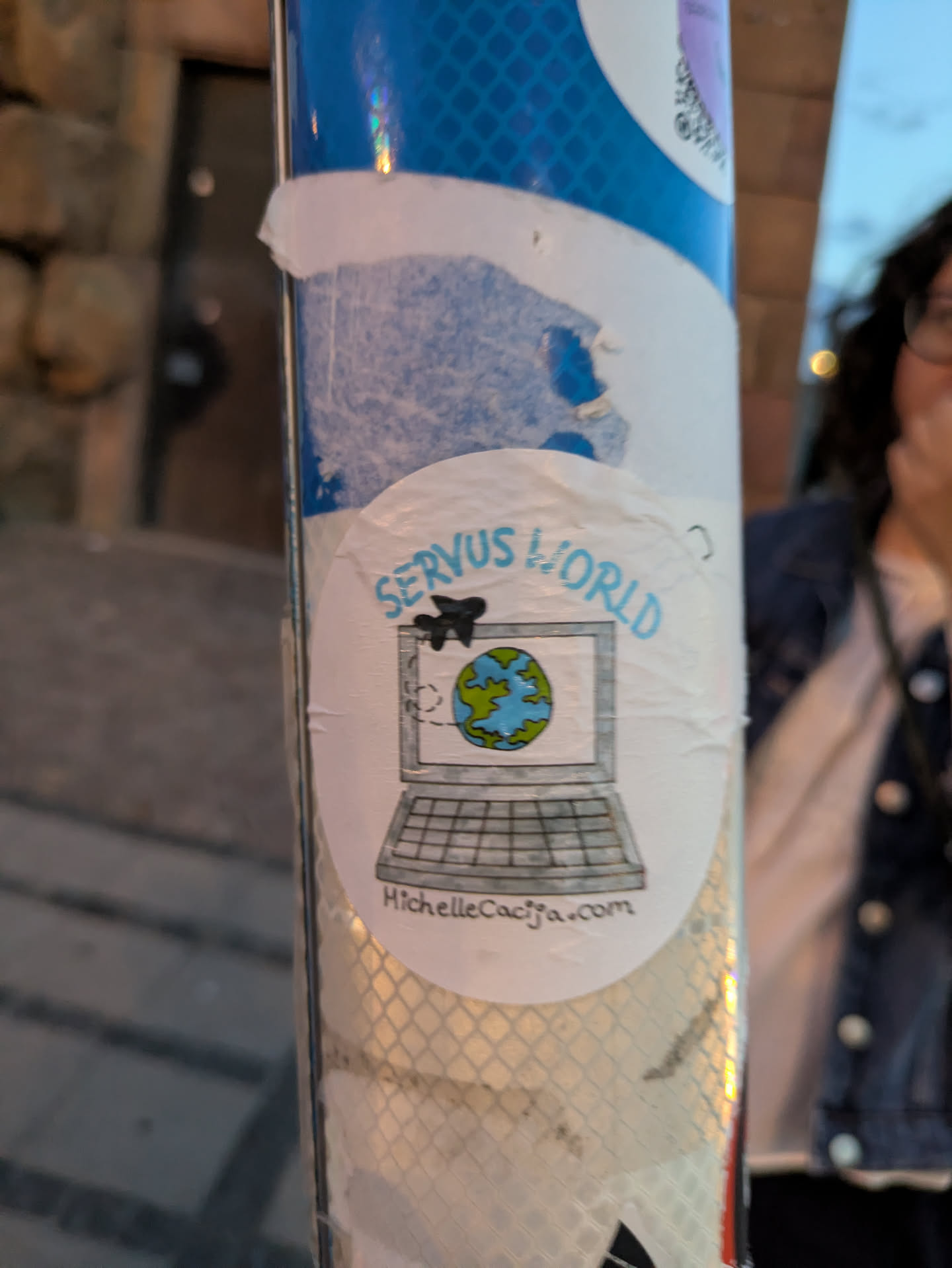 A circular sticker features a hand-drawn illustration of an open laptop with a screen displaying a globe. A small airplane is depicted flying from the top left of the screen towards the globe. The text 'SERVUS WORLD' is written in a light blue, slightly whimsical font above the laptop. Below the laptop screen, the website address 'michellecacifa.com' is written in a smaller, darker font. The sticker appears to be made of a white material with a slightly textured or worn appearance.