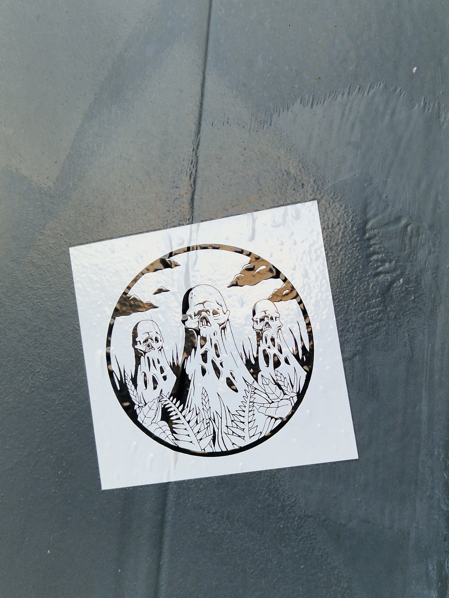 The sticker displays a circular design with stark black line art on a white background, affixed to a dark, textured surface. The central motif consists of three grotesque, skull-like figures that appear to be melting or dripping, with elongated, organic shapes flowing from them. These figures are intertwined with intricate botanical elements, resembling leaves and ferns, suggesting an eerie blend of life and decay. The background within the circular frame shows subtle cloud formations. The overall aesthetic is dark, macabre, and illustrative, evoking themes of gothic art or dark fantasy.