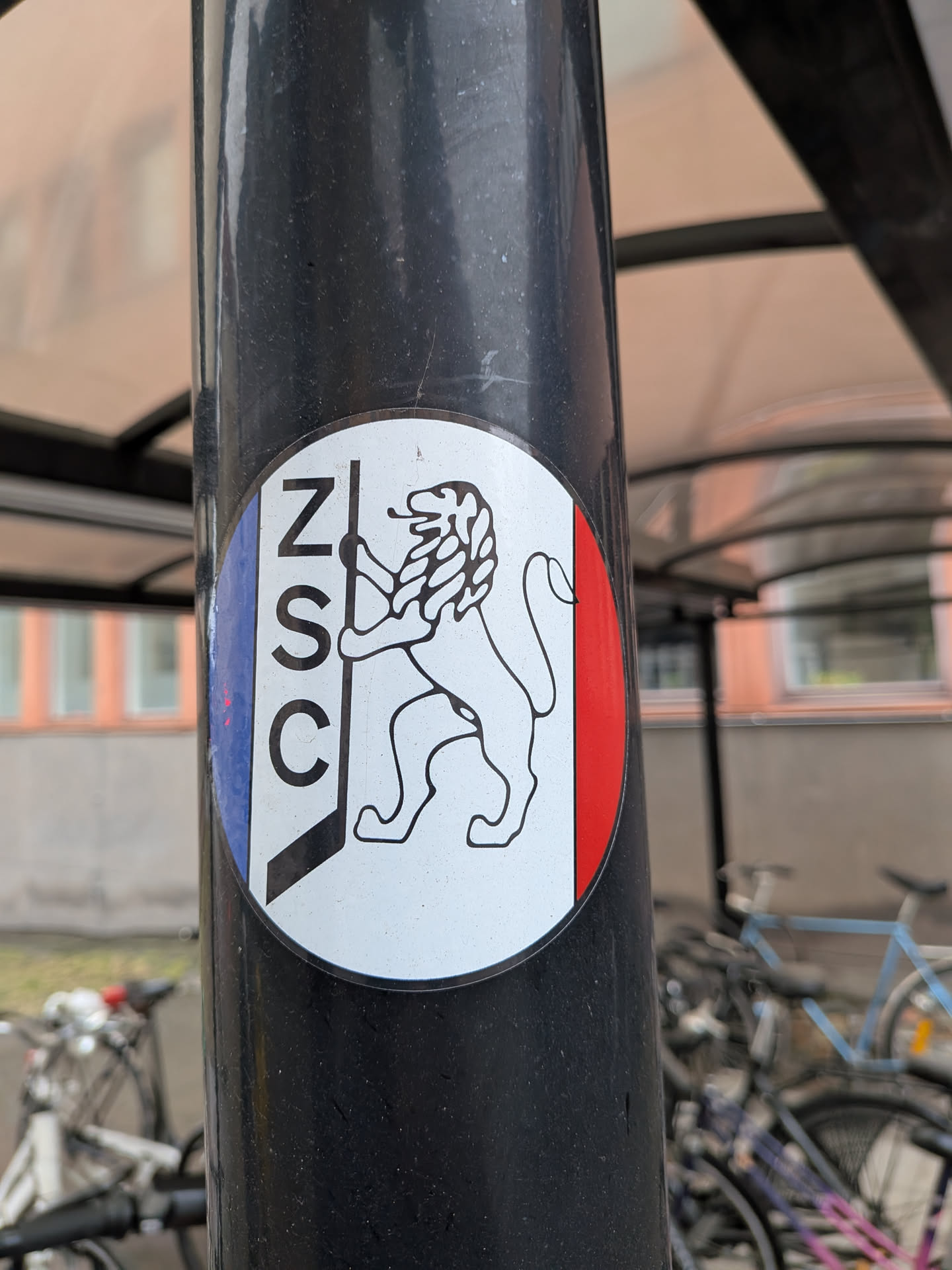 The sticker is circular, predominantly white in the center, with vertical blue and red stripes on its left and right edges, respectively, reminiscent of the French flag. In the center, there is a black outline drawing of a lion rampant, standing on its hind legs and facing right. The lion appears to be holding a vertical stick or pole with its front paws. To the left of the lion, vertically aligned within the blue stripe and extending into the white area, are the black capital letters 'ZSC'. The sticker is applied to a dark grey or black pole and shows minor signs of wear.