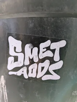 A close-up of a sticker with the text "SMET LADS" in a stylized, rounded font. The sticker is black with white lettering, affixed to a dark, possibly metallic or plastic, surface.