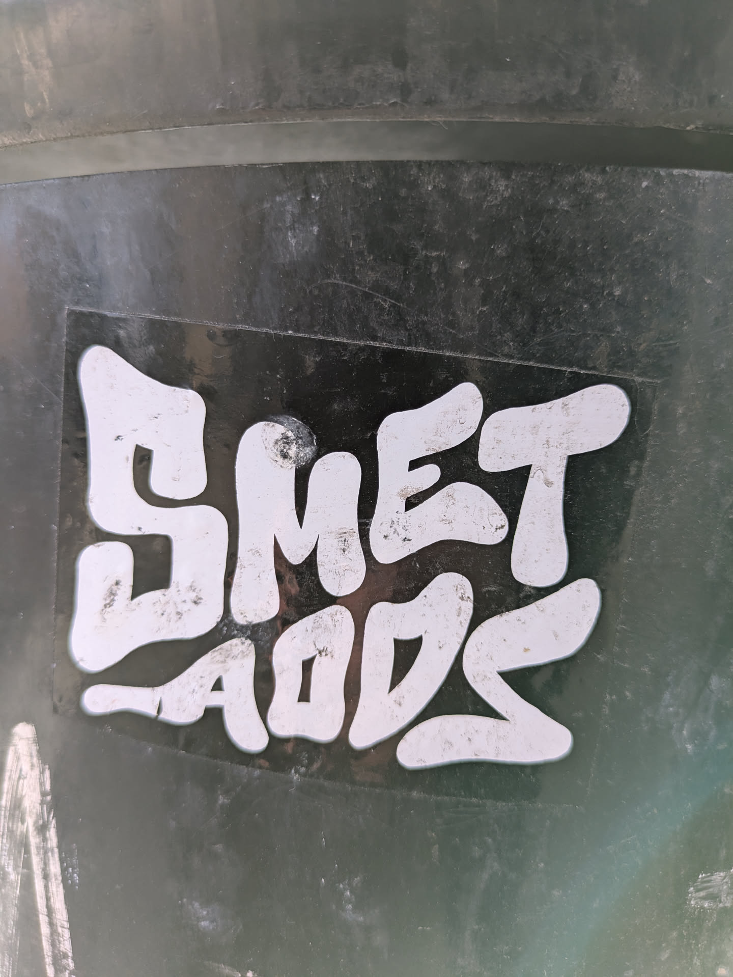 A close-up of a sticker with the text "SMET LADS" in a stylized, rounded font. The sticker is black with white lettering, affixed to a dark, possibly metallic or plastic, surface.