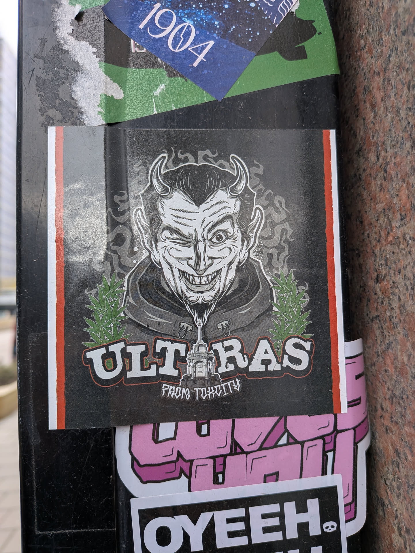 The central sticker features a stylized, cartoonish depiction of a winking devil with horns, pointed ears, and a sharp-toothed grin. The devil wears a hooded garment, and a liquid-like substance drips from its chin, forming a detailed architectural structure resembling a city building or monument. On either side of this central image are green cannabis leaves. The word "ULTRAS" is prominently displayed in large, white, block letters with a black outline and a subtle red inner shadow, positioned above the architectural element. Below "ULTRAS" and the building, in smaller, stylized white text, it reads "FROM TOXCITY". The main design is set against a dark grey background with a smoky or cloudy pattern, and the entire sticker is bordered on the left and right by thin vertical stripes of red and white.